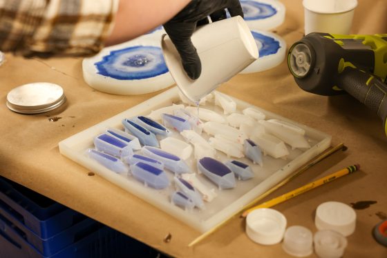 A close-up of a hand in a black glove pouring clear resin from a white cup over a silicone mold containing small, faceted, crystal-shaped pieces of blue and white resin.