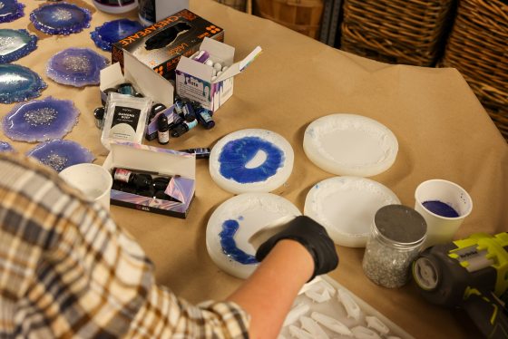 A close-up, overhead shot of a person wearing a black glove applying blue resin/pigment to a coaster mold. The table is covered with resin supplies, including color bottles, glitter, and other coaster molds.