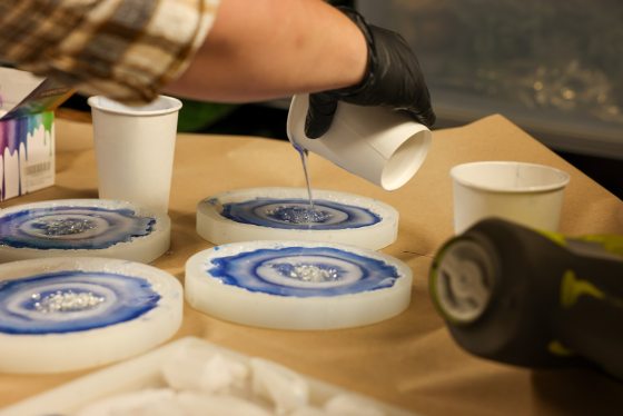 A person in a black glove pours clear resin from a small paper cup into the center of a blue and white geode coaster mold. Three other filled coasters are visible on the brown work surface.