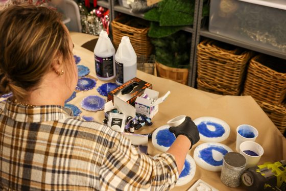 A woman with her back to the camera, wearing a plaid shirt and black gloves, works on a craft project involving pouring resin into circular molds on a table, with several finished blue and purple geode pieces drying in the background.