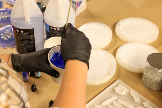 A close-up of a hand in a black glove holding a paper cup filled with vibrant blue colored resin, stirring it with a wooden stick over a work surface with empty coaster molds.