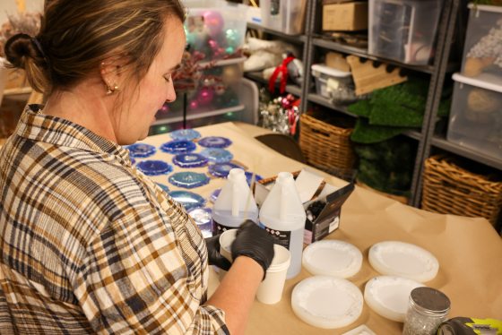 A side view of a woman in a plaid shirt and black gloves preparing resin mix next to large bottles of resin and several empty circular coaster molds on a table. Finished geode pieces are visible in the background.