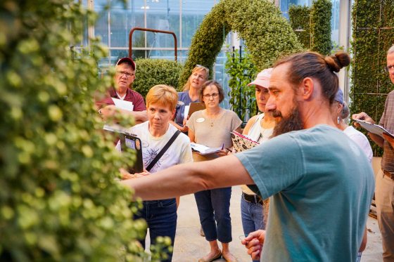 A gardening instructor with a beard and hair in a bun gestures while leading a small group of adults on a focused tour or class in a greenhouse.