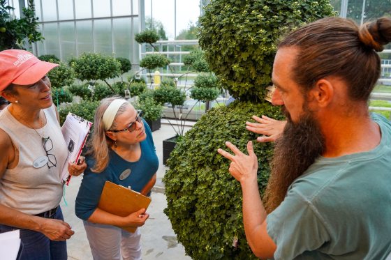 Gardening expert instructing two students on topiary care and shaping techniques inside a large glass-walled greenhouse.