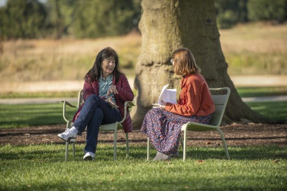 Joy Harjo sitting outdoors in a green metal chair, gesturing while speaking to a woman across from her who is holding a notebook. They are on a green lawn with a large tree trunk visible in the background.