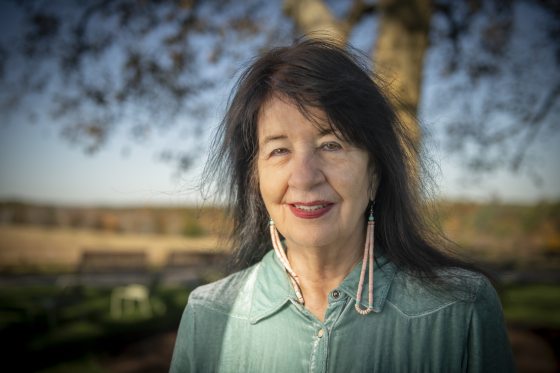 Outdoor portrait of poet Joy Harjo smiling at the camera, wearing a light teal velvet collared shirt and long beaded earrings, with a large tree and clear blue sky out of focus behind her.