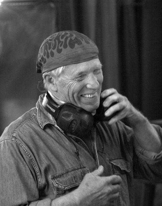 Black and white portrait of sculptor André Harvey smiling, wearing a denim shirt and a bandana with a flame pattern, with noise-canceling headphones resting around his neck.
