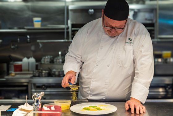 A person in a white chef coat, in a kitchen, sprinkling yellow powder onto a plate of food. 