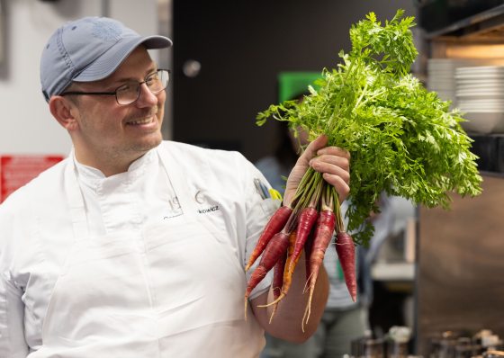 A smiling chef in a white coat, apron, and baseball cap holds up a large bunch of fresh, vibrant red carrots with their green tops still attached.