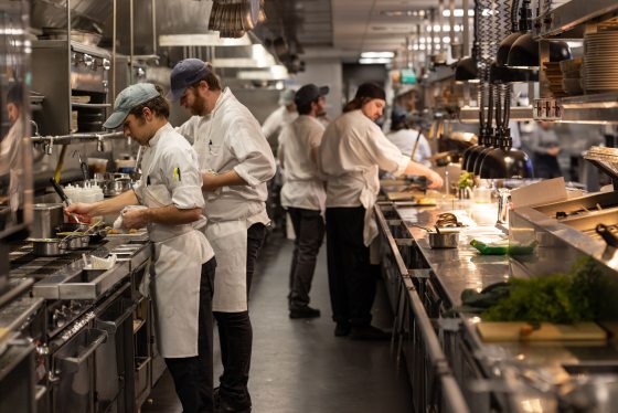 A busy professional kitchen scene with several chefs in white coats and aprons working along a long, stainless steel counter, preparing and plating food.