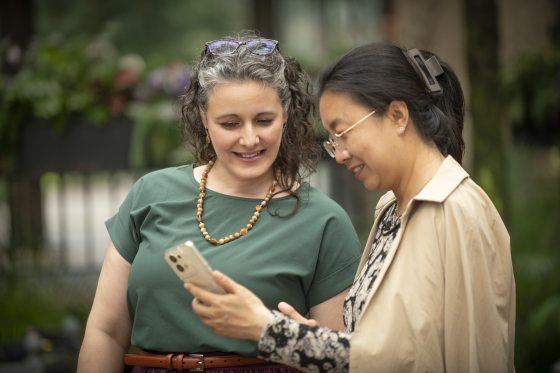 Two women, one in a green top and the other in a beige coat, looking together at content on a smartphone screen.
