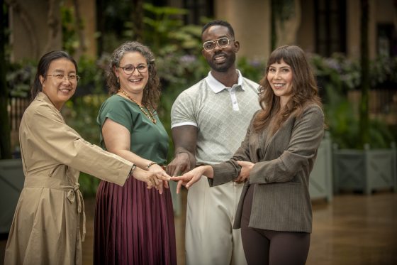A group portrait of four individuals—two women and two men—smiling and interlocking their hands in the center, taken indoors in a garden or conservatory setting.