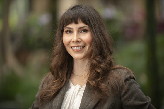 Close-up smiling headshot of a woman with dark hair, bangs, and a brown pinstripe blazer.