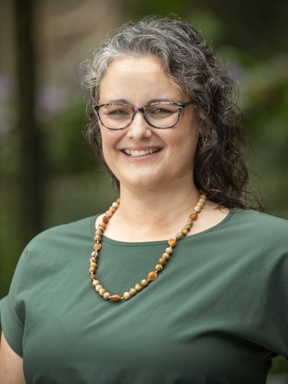 Close-up smiling headshot of a woman with gray, curly hair, glasses, a green shirt, and a beaded amber necklace.
