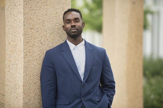 A man in a navy blue suit and white shirt posing outdoors, leaning against a tan architectural column.