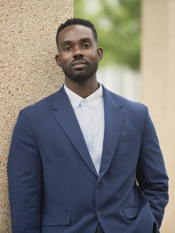 A man in a navy blue suit and white shirt posing outdoors, leaning against a tan architectural column.