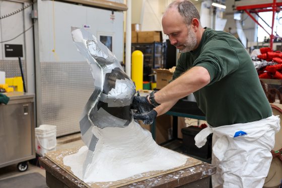 A man in a workshop carefully applies tape and foil to a dark, faceted sculpture model, preparing the surface for the molding process.
