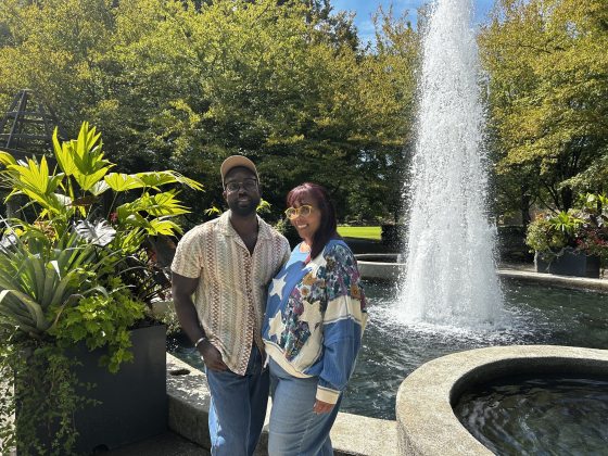 Fellow Nathaniel Cody and Marylynn Mack, Executive Director of Memphis Botanical Garden, standing and smiling in front of a tall, spouting water fountain and large potted plants.