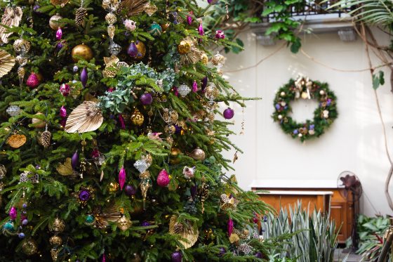 A close-up view of a dense Christmas tree decorated with a mix of colorful, ornate ornaments in shades of purple, gold, and magenta, alongside natural elements like pinecones and dried leaves, with a simple wreath visible on a white wall in the background.