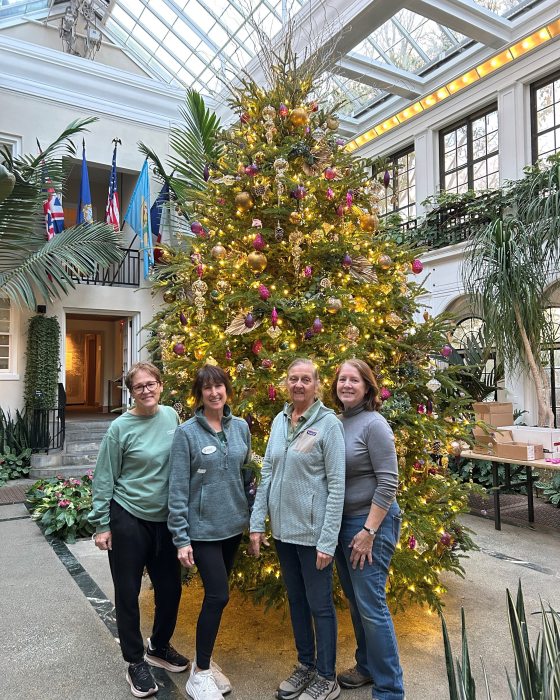 Four women stand together smiling in front of a massive, brightly lit and decorated Christmas tree inside a sunlit glass conservatory or atrium.