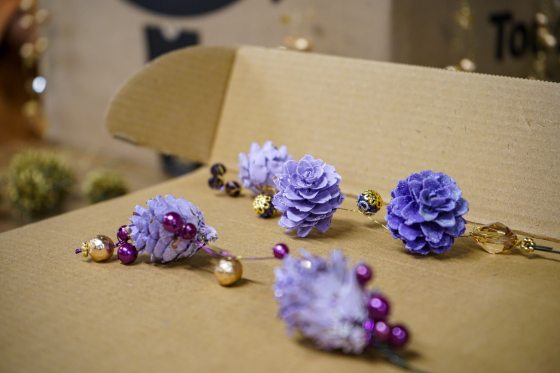 A close-up of decorative elements, including purple bead-like accents and lavender-colored painted pinecones, arranged on a piece of cardboard.