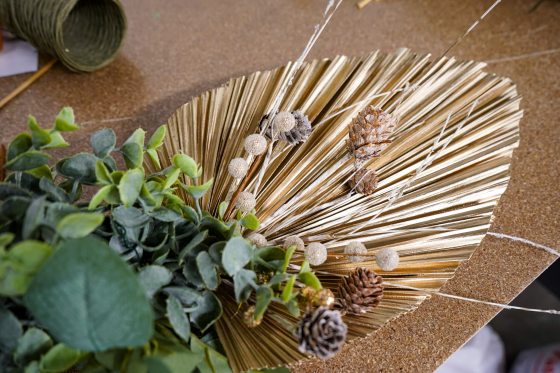  A close-up of a holiday decoration component being assembled, featuring a large, metallic gold, pleated, fan-shaped palm leaf, adorned with sprigs of faux eucalyptus leaves, small pinecones, and delicate white glittery spheres on thin wires.