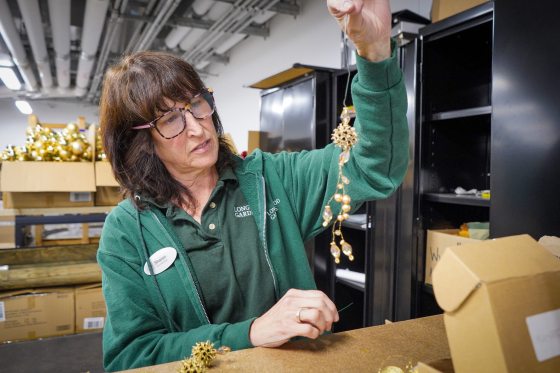 A woman in a green shirt and glasses holds up a delicate gold and pearl-like decorative ornament, seemingly for a Christmas tree, while seated at a workbench in a storage area.