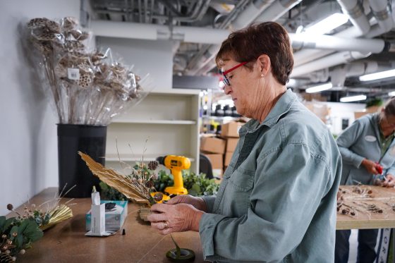 A woman in a green shirt and glasses works on creating a decorative piece, possibly for a Christmas display, using dried natural materials and glue, with other supplies and a drill visible on the counter behind her.