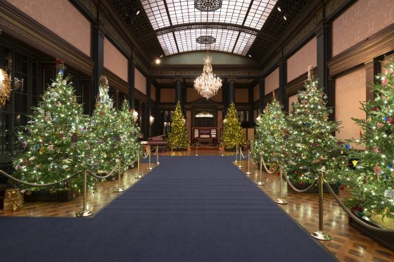 A grand hall with a high, vaulted glass ceiling and a large chandelier. A wide, dark blue carpet runs down the center aisle, flanked by rows of heavily decorated Christmas trees on either side.