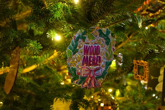 A close-up of a highly detailed, glittery Christmas ornament shaped like a wreath or medallion. The ornament is primarily green and gold with a pink bow and the words "WOOD MERE!" in the center, hanging from a Christmas tree branch.