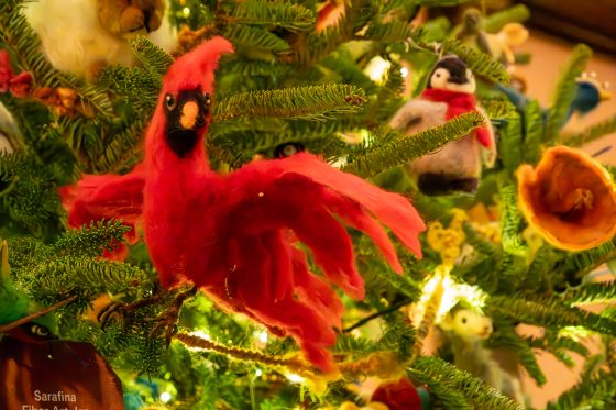 A close-up photo of a Christmas tree branch featuring a striking, large ornament shaped like a red cardinal, made from needle-felted fiber art. A smaller felt penguin ornament is visible next to it.