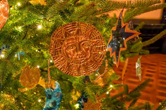 A close-up of a Christmas tree decorated with a prominent, round, pressed copper or bronze ornament featuring a smiling sun face surrounded by rays. A reflective blue star ornament is visible to the right.
