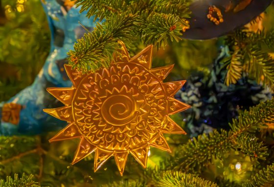 A close-up of a golden, star-shaped ornament with pointed rays and an intricate sunburst or spiral pattern etched into the center, hanging on a brightly lit Christmas tree.