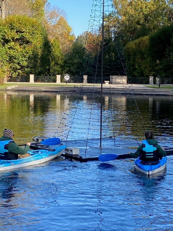 Two people in blue kayaks on a lake working to position a large, cone-shaped light structure made of netting and string lights.