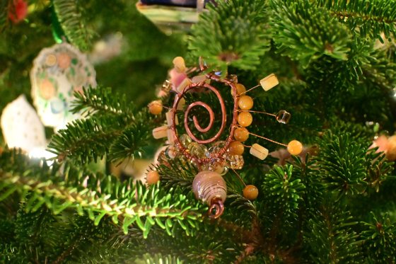 A unique Christmas ornament made of twisted copper wire forming a spiral, adorned with orange and tan beads and small wire accents, hanging on a green Christmas tree.