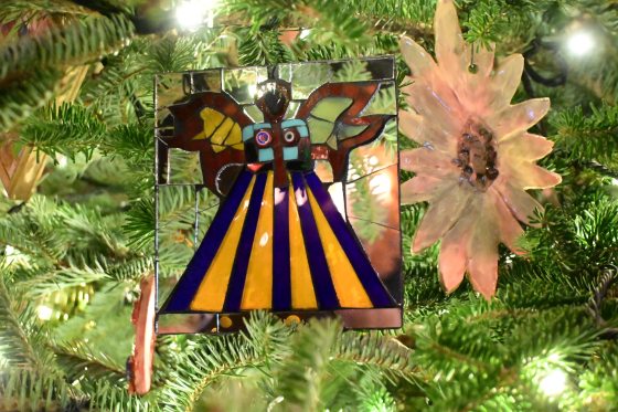 A Christmas tree ornament made of stained glass pieces in the shape of an abstract angel with a yellow and purple striped skirt. To the right, there is a lighter, translucent flower-shaped ornament.