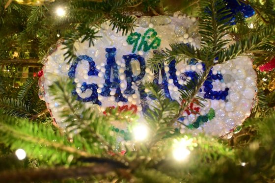 A close-up of a Christmas tree ornament decorated with clear and blue glass beads that spell out "The Garage" in a festive, multi-colored style. The ornament is nestled among the green tree branches.