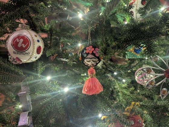 A close-up of several ornaments on a Christmas tree. One is a white glass ball with a red logo and the word "EL FUTURO." Another is a round, felted ornament with a white figure and small pink flowers, featuring a pink yarn tassel hanging below it.
