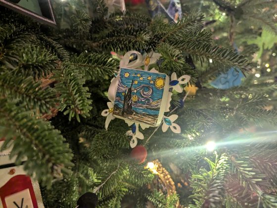 A small square ornament hangs on a Christmas tree. It features a miniature rendition of Vincent van Gogh's The Starry Night painting, surrounded by white, flower-shaped plastic pieces.