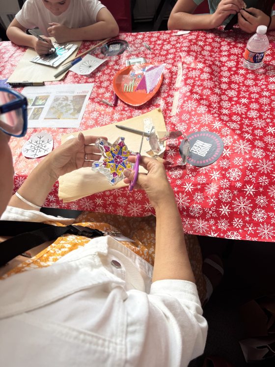 An overhead view of a person cutting out a snowflake-shaped piece from a clear plastic sheet using purple scissors. The table is covered with a red and white snowflake-patterned tablecloth, and crafting supplies are visible.