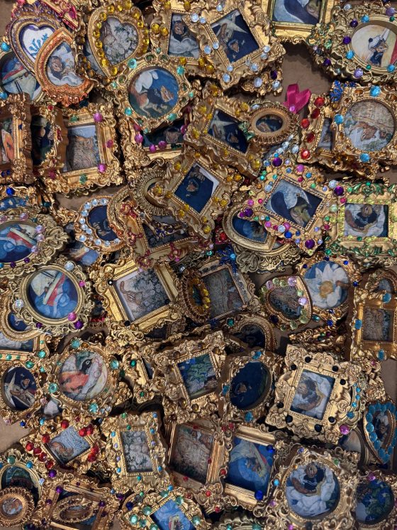 A full frame close-up of a large pile of intricate, small, gold-colored picture frames, each holding a miniature picture and heavily embellished with colorful glass gems or jewels.