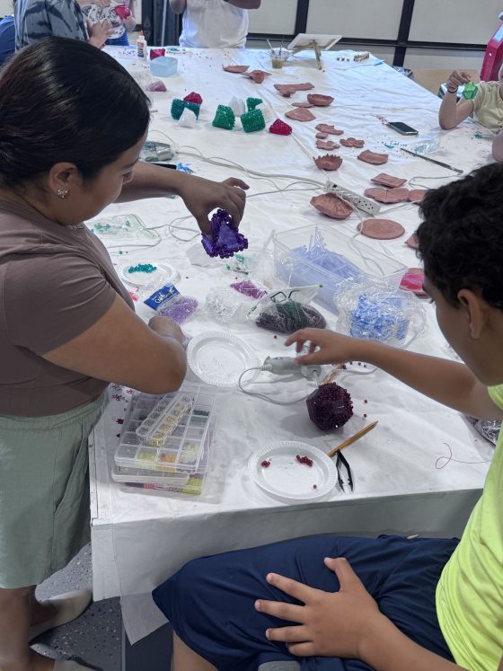 A person is using a hot glue gun to attach colorful plastic beads to a purple, rough-textured shape at a large table. Various crafting supplies and unfinished beaded or molded clay shapes are spread across the table.