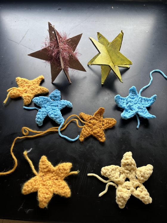 An arrangement of handmade star-shaped crafts on a black surface. The pieces include three-dimensional stars made from cardboard/paper, one brown with pink tinsel and one painted yellow-green, alongside several flat, crocheted star-shaped ornaments in yellow, blue, and cream.