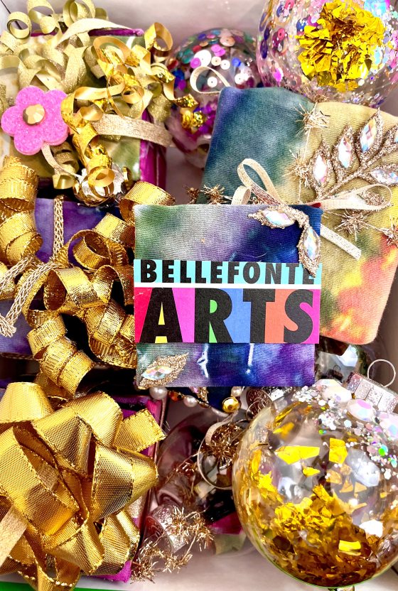 A bright, close-up shot of a box filled with colorful Christmas ornaments and decorations. The focus is on a central card that reads "BELLEFONTE ARTS" in colorful, block letters over a tie-dye background, surrounded by gold ribbon bows, sequined globe ornaments, and small, wrapped tie-dye gift boxes.