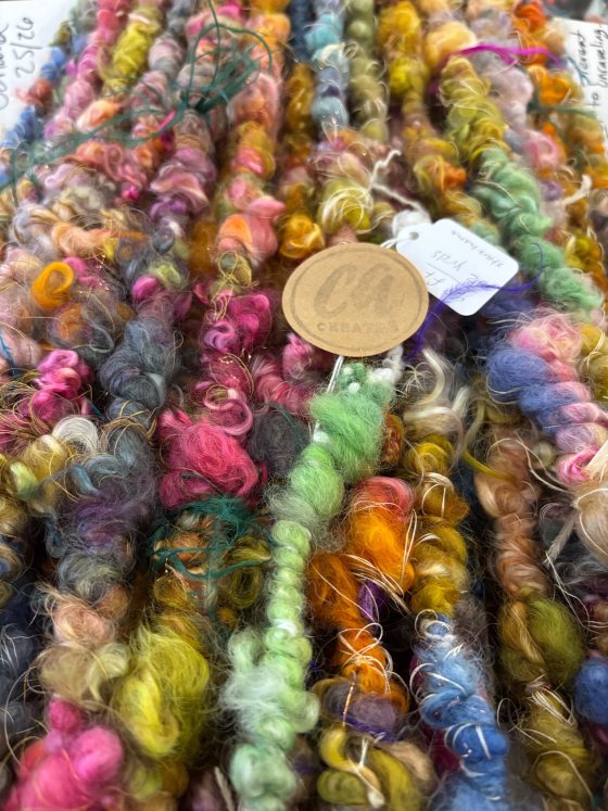 A close-up, high-texture shot of various colorful art yarns or fibers, including shades of pink, green, yellow, and blue, twisted into locks. A small circular tag with the letters "CA" is visible in the center.