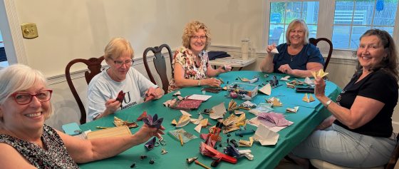 Five older women sitting around a green-covered table indoors, smiling at the camera while working on paper folding/origami crafts, including multi-pointed stars.