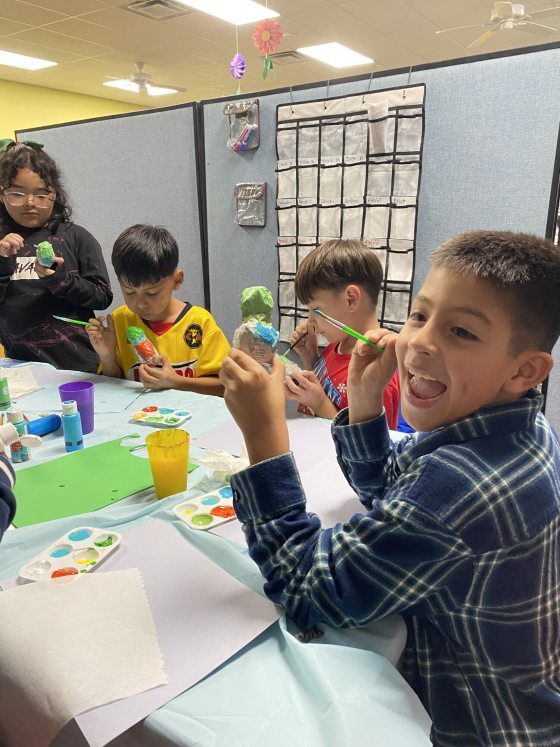 A close-up of several children sitting at a table, actively and happily painting small ceramic or papier-mâché crafts. The boy in the foreground smiles brightly at the camera while painting a blue and white figure.