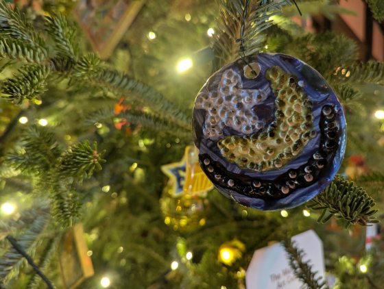 A close-up of a round, ceramic or clay ornament hanging on a Christmas tree. The ornament is glazed in dark blue with a raised, textured design in white and gold/yellow, resembling a crescent moon or abstract celestial body.