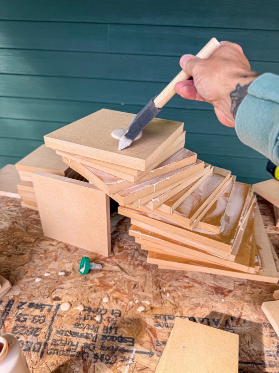 A close-up shows a hand using a foam brush to apply white glue to a stack of square MDF boards, illustrating the early assembly stage of the sculpture's core.
