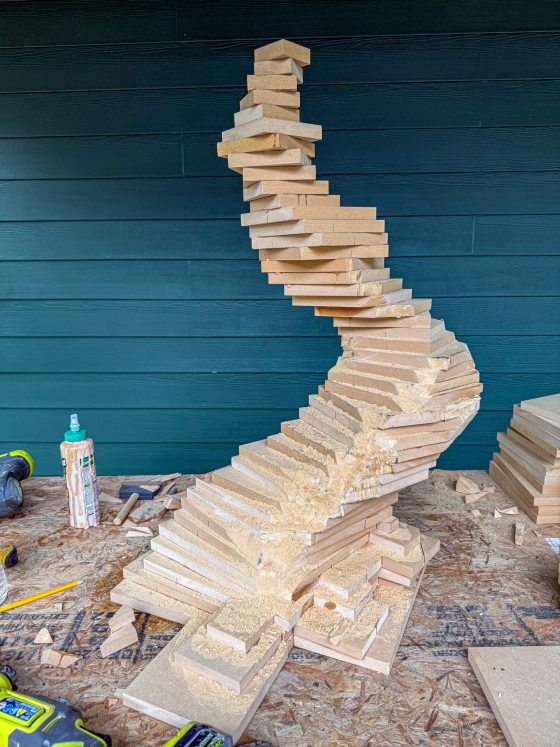A tall stack of square MDF boards is glued together in a curved, spiraling form to create the initial rough shape of the bonsai tree trunk.
